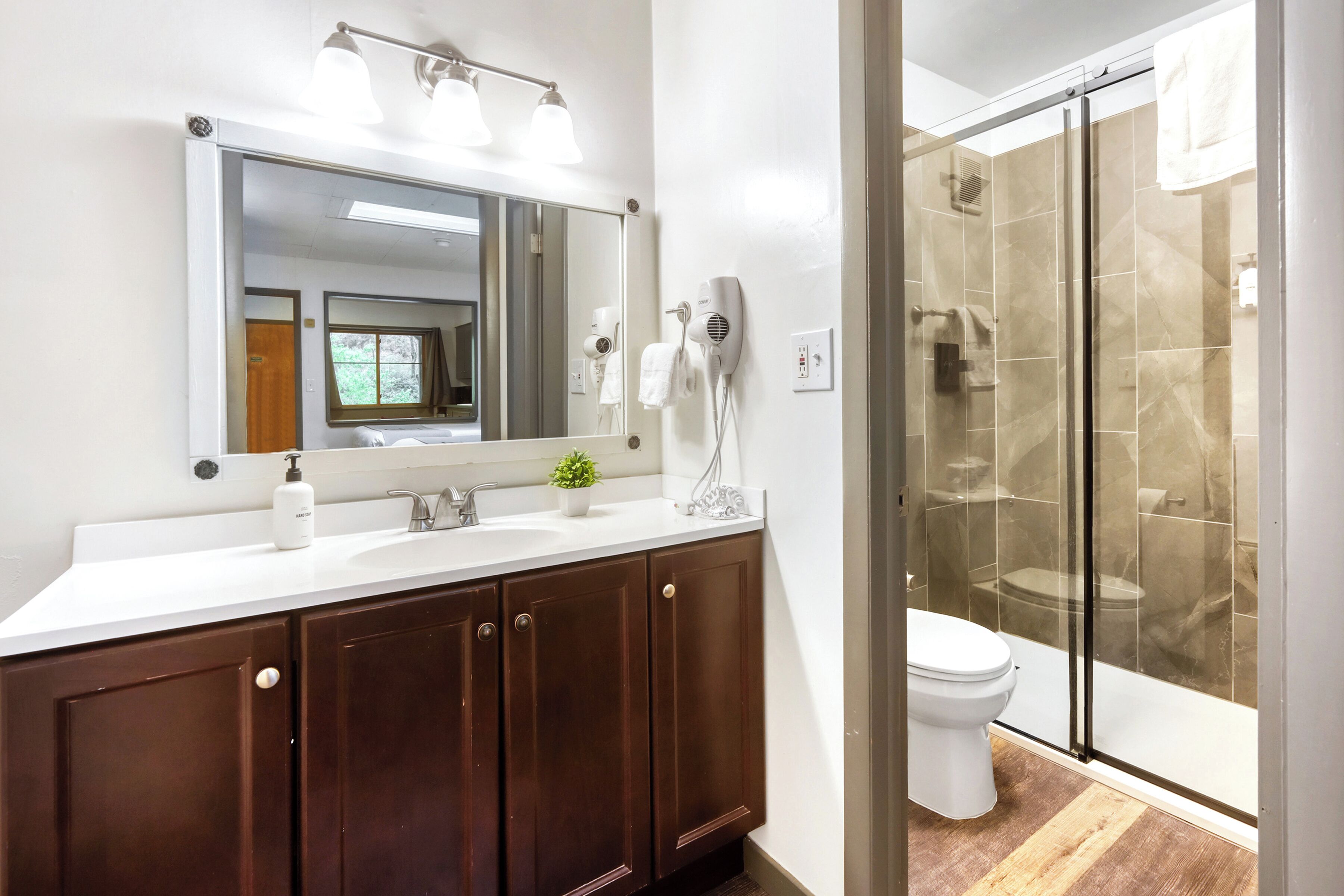 A bathroom with a white sink, dark wooden cabinets, a large mirror, a handheld hair dryer, and a glass-enclosed shower.