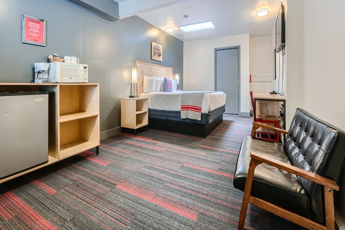 Une chambre d’hôtel comprenant un lit, un bureau avec un micro-ondes, un réfrigérateur, une chaise et une petite table sur laquelle se trouve un panier rempli de divers objets.