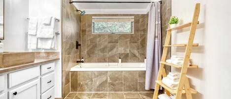 Une salle de bain moderne avec une baignoire, une étagère en bois où sont posées des serviettes et une fenêtre donnant sur des arbres.