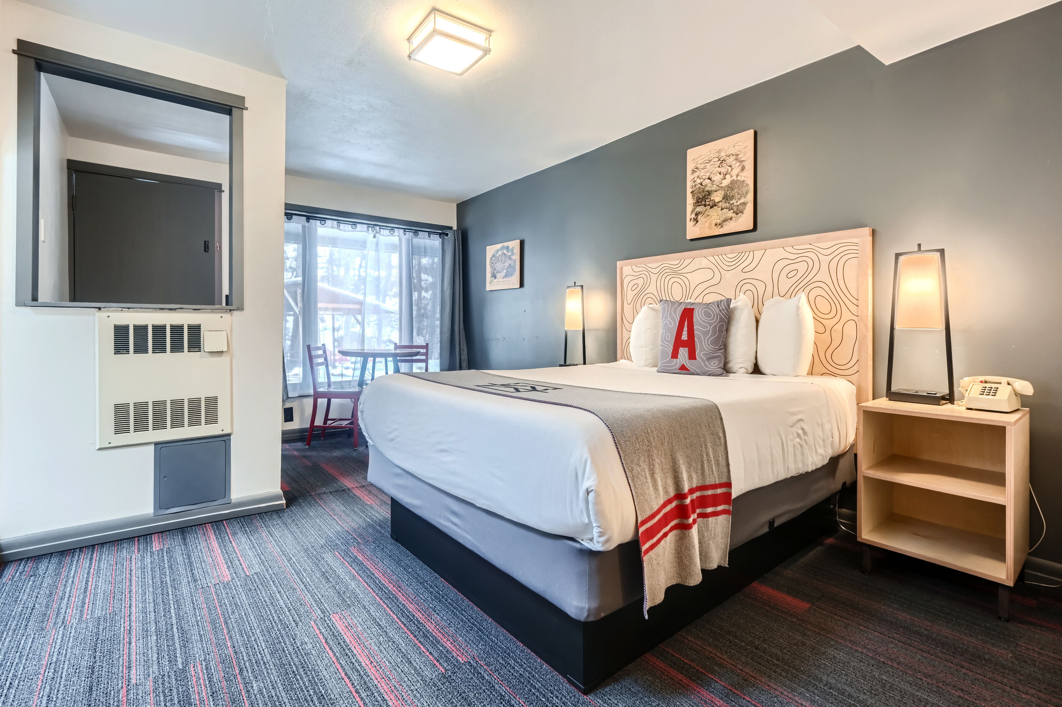 A hotel room with a large bed, a flat-screen TV mounted on the wall, a bedside table with a lamp, and a window with curtains.