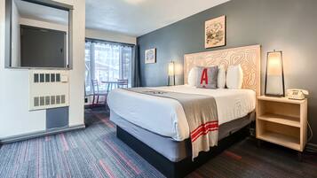 A hotel room with a large bed, a flat-screen TV mounted on the wall, a bedside table with a lamp, and a window with curtains.