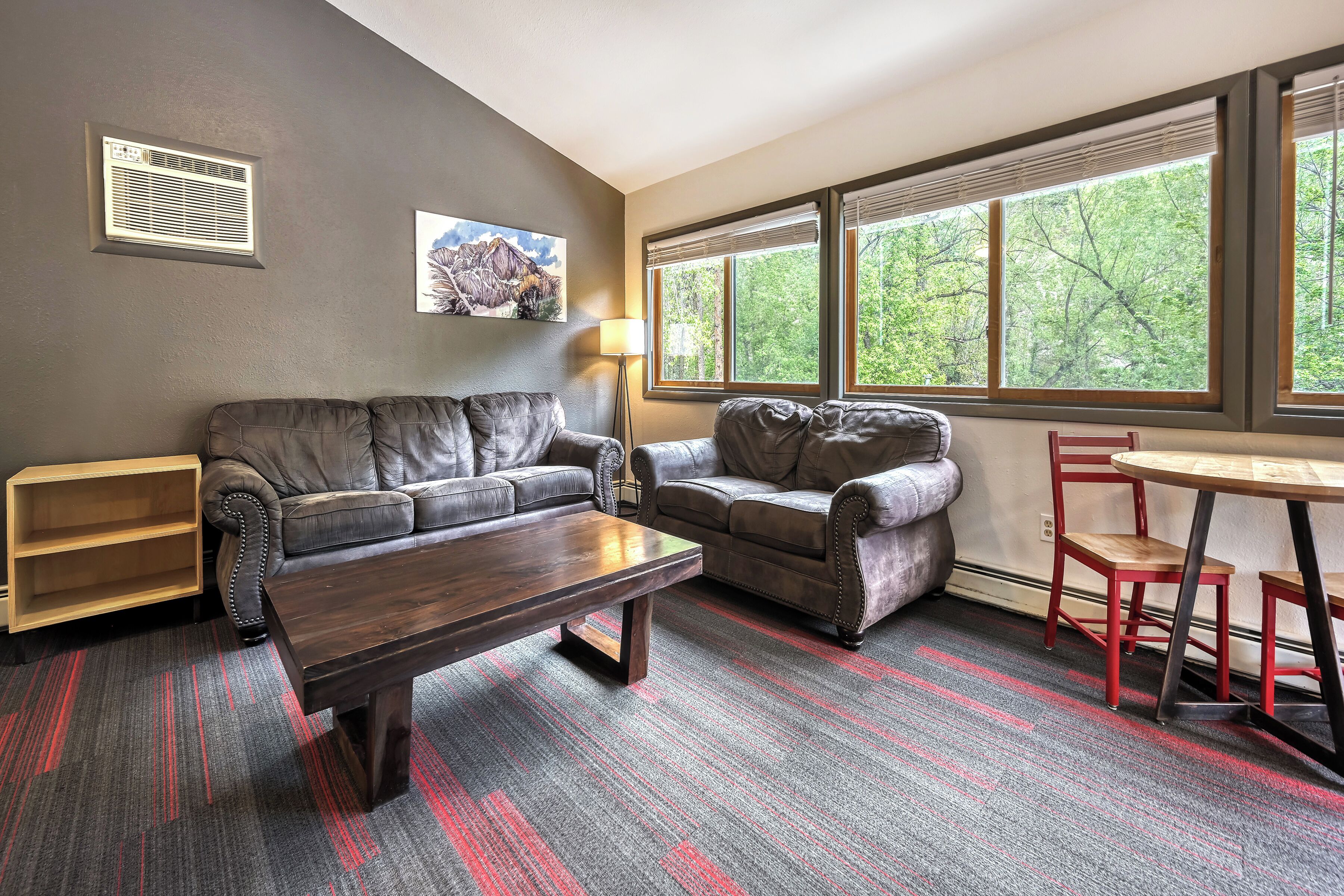 A living room with a sofa, a coffee table, a lamp, a red chair, and a window with a view of trees.