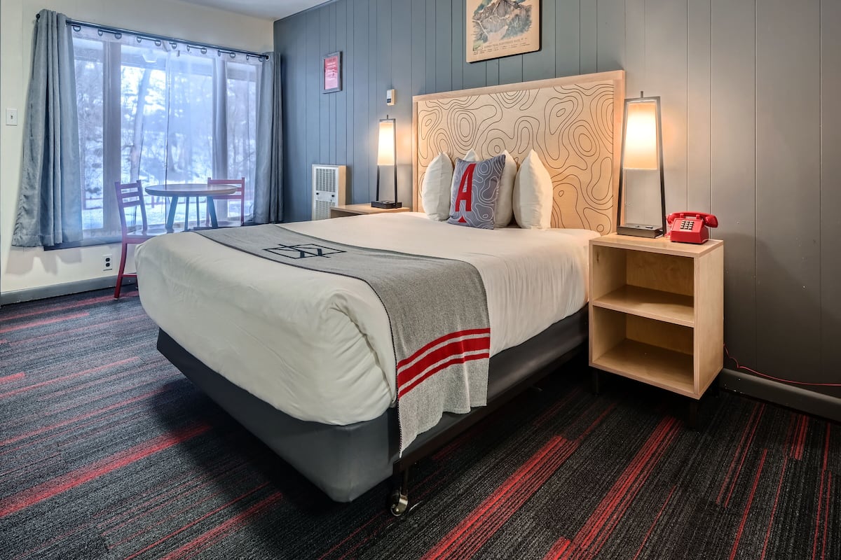 Une chambre d’hôtel avec un grand lit, une table de chevet avec une lampe, un téléphone rouge et une vue sur l’extérieur par une fenêtre.