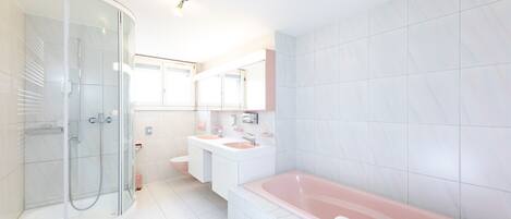 Un baño moderno con ducha envuelta en vidrio, una bañera rosa grande y elementos blancos.