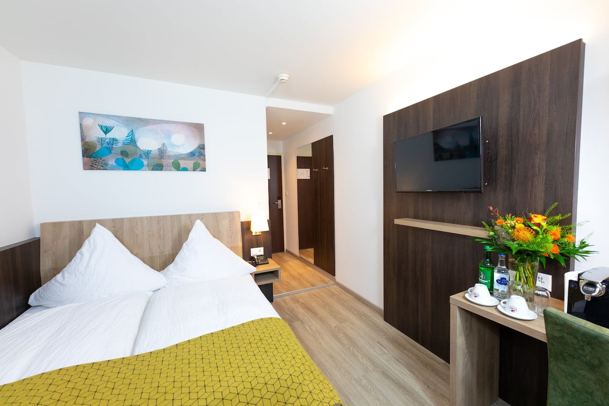 A hotel room with a bed, a desk with a flat-screen TV, a painting on the wall, and a flower arrangement.