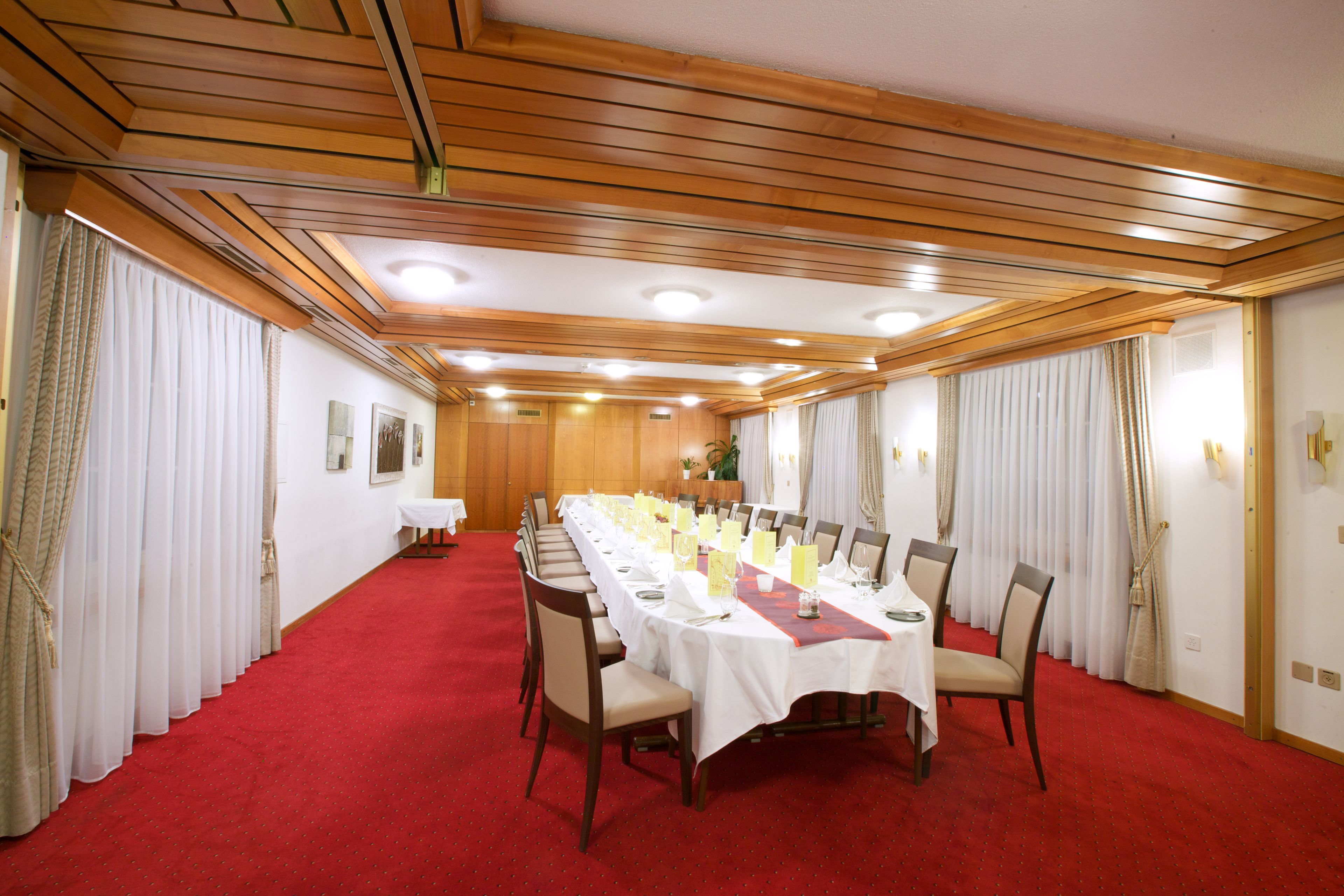 A conference room with a long table set for a meeting, wooden panel ceiling, and red carpet flooring.