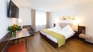 A hotel room with a bed, a desk, a chair, a TV, and a window with curtains. - Alfa Hotel (Birsfelden)