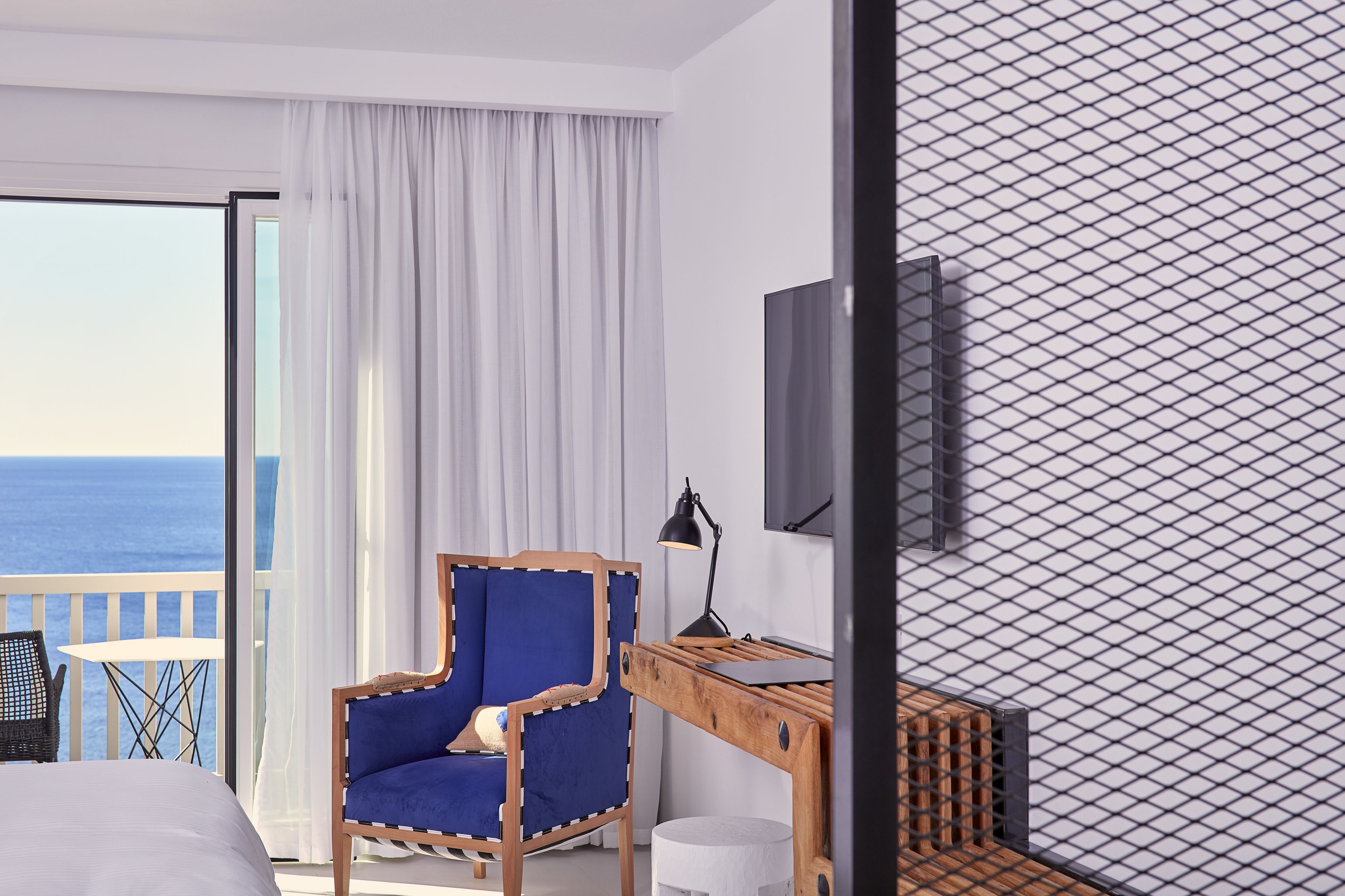Une chambre d’hôtel avec un grand lit, un fauteuil bleu, un bureau en bois, un téléviseur fixé au mur et une vue sur la mer par la fenêtre.