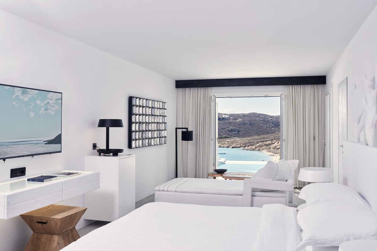 A modern hotel room with a large bed, a desk with a computer, a chair, and a view of the sea.