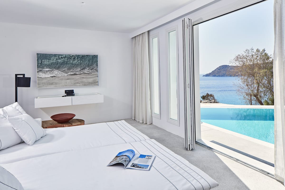 Ett modernt hotellrum med en stor säng, utsikt över havet, en swimmingpool och en matplats med en röd skål.