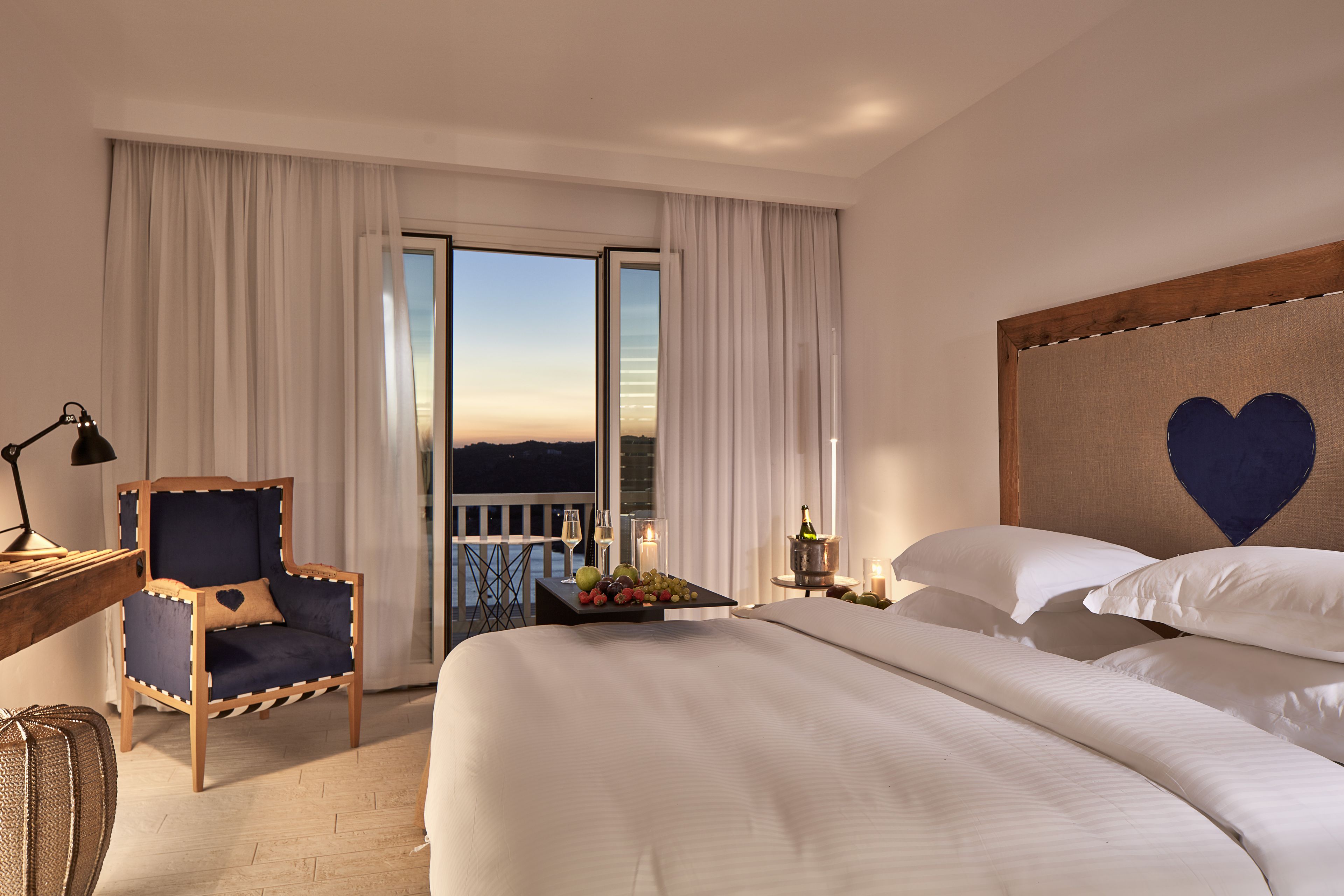 Une chambre d’hôtel avec un grand lit, une chaise, un bureau avec une lampe, une table de chevet avec des bougies et une vue sur le ciel par la fenêtre.