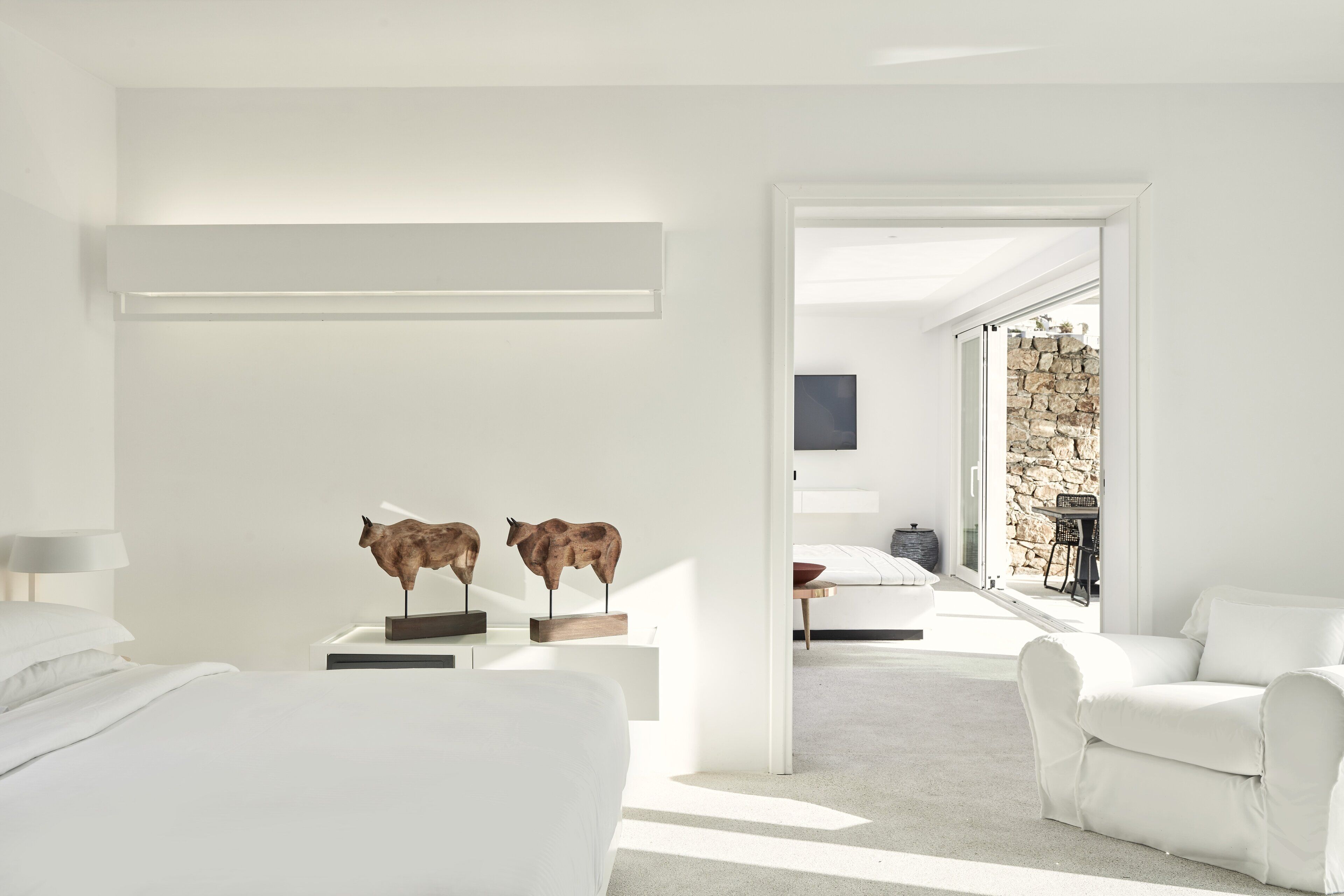 A modern hotel room with a minimalist design, featuring a bed, a white armchair, and a desk with two wooden sculptures.