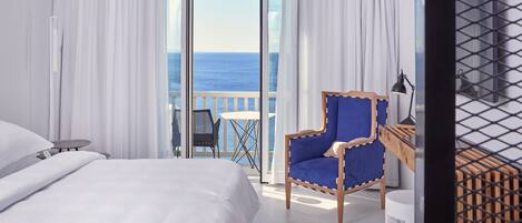 Une chambre d’hôtel avec un lit, un fauteuil bleu, une petite table et une vue sur la mer, visible à travers une porte vitrée.