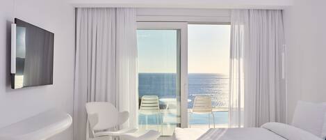A modern hotel room with a large bed, a sitting area with two chairs, a small table, a bathtub, and a TV. The room has a large window with a view of the ocean.