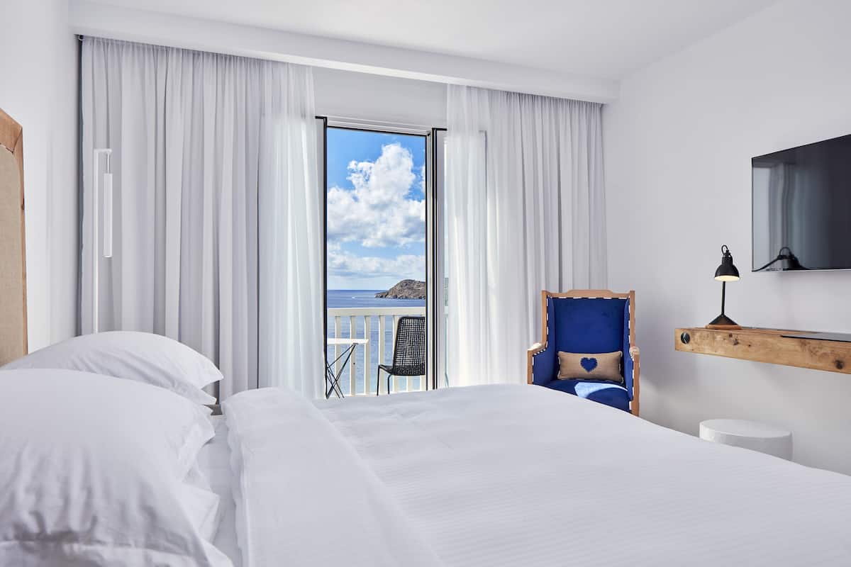 A hotel room with a large bed, a chair, a small table, a view of the sea, and a mounted TV.