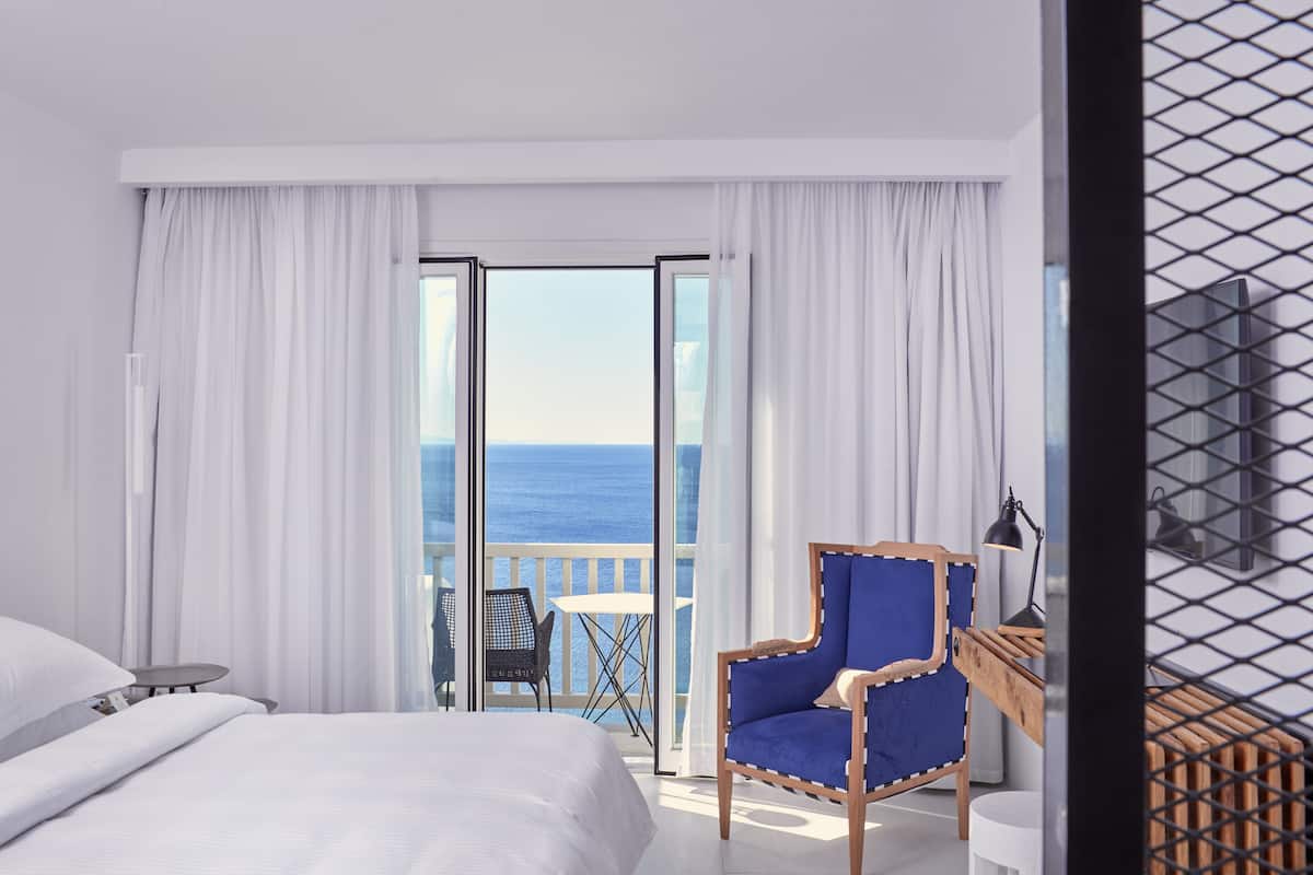 A hotel room with a bed, a blue armchair, a small table, and a view of the sea through large windows.