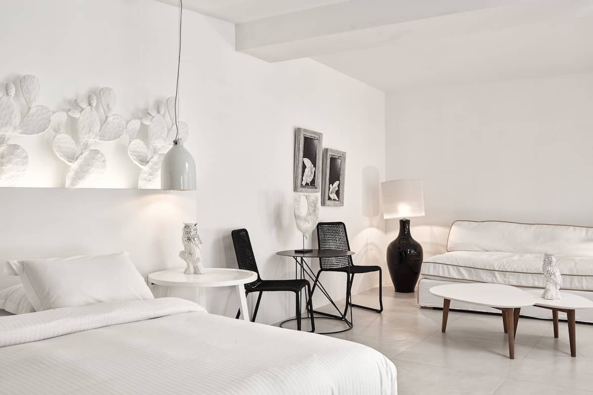 A modern hotel room with a large bed, a round dining table, two black chairs, a white sofa, and a decorative cactus wall art.
