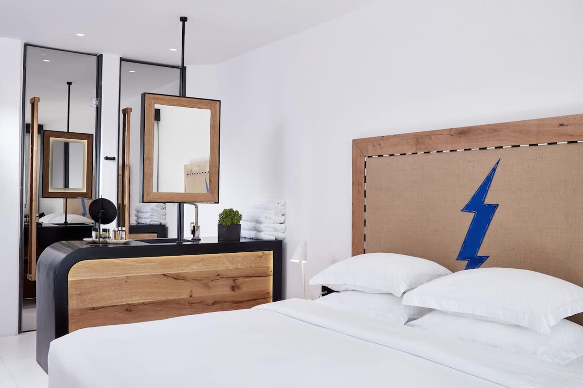 A modern bedroom with a wooden headboard, a large mirror, a bedside lamp, and a bed with white bedding.