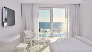 Una habitación de hotel moderna con una cama grande, una zona de estar con dos sillones, una mesita pequeña, una bañera y un ventanal con vistas al océano.