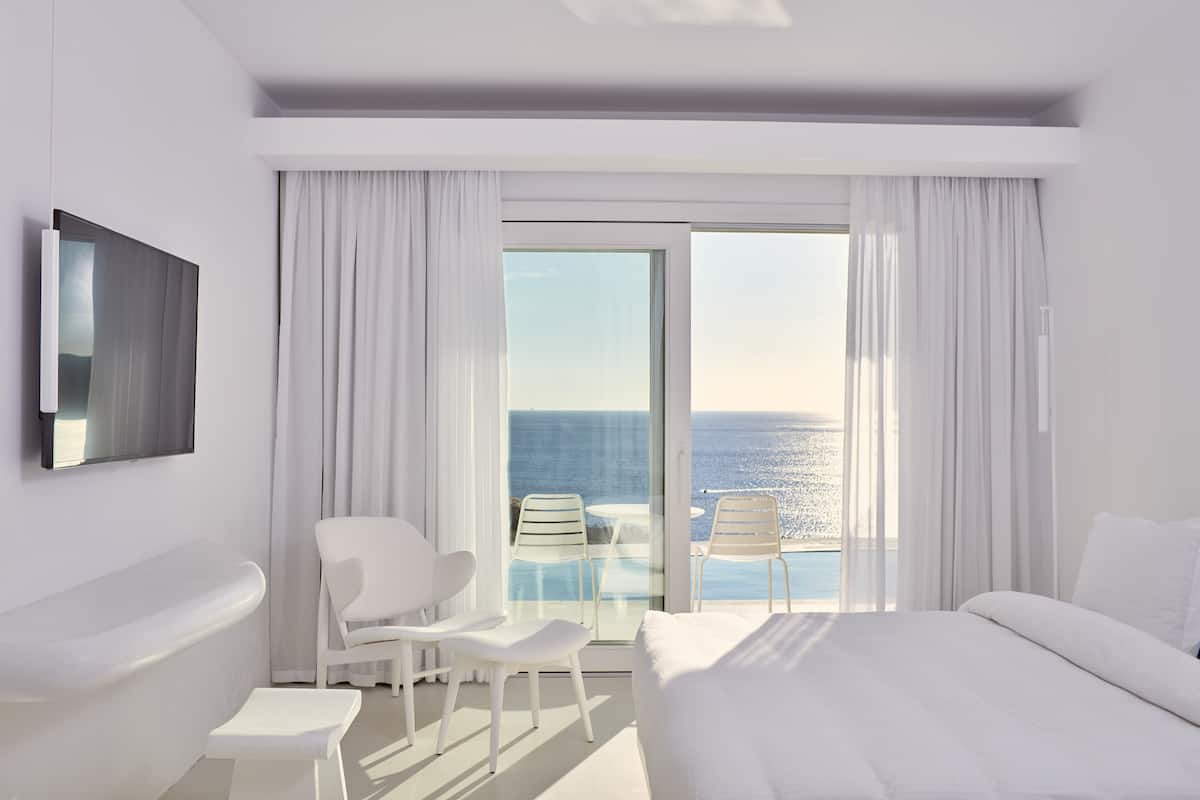 A modern hotel room with a large bed, a sitting area with two chairs, a small table, a bathtub, and a large window with a view of the ocean.