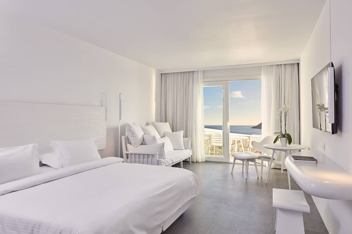 A modern hotel room with a large bed, a sofa, a dining area, and a balcony with a view of the sea.
