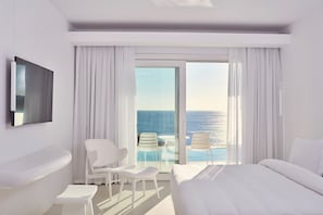 Une chambre d’hôtel comprenant un lit, une télévision, une petite table, deux chaises, une salle de bain et un balcon avec vue sur la mer.