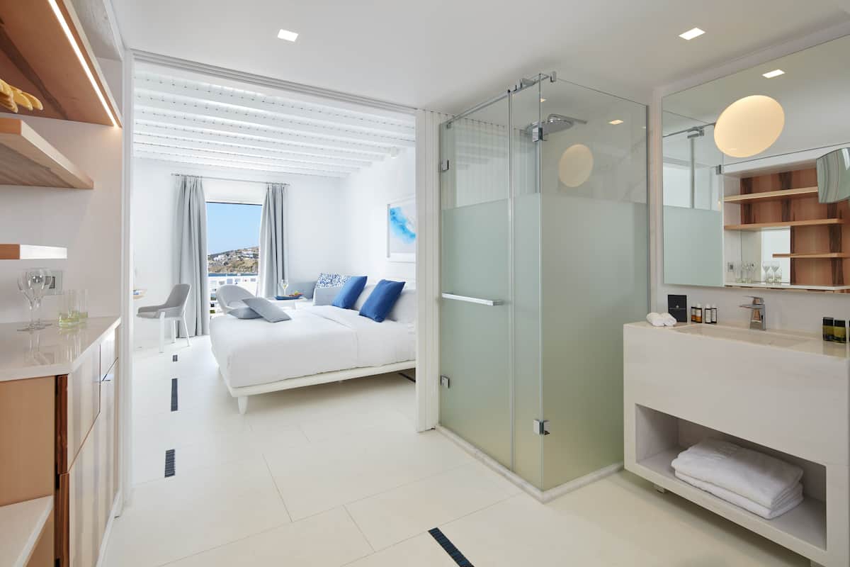 Een moderne hotelkamer met een bed, een douche in een glazen cabine, een grote spiegel en uitzicht op de stad.