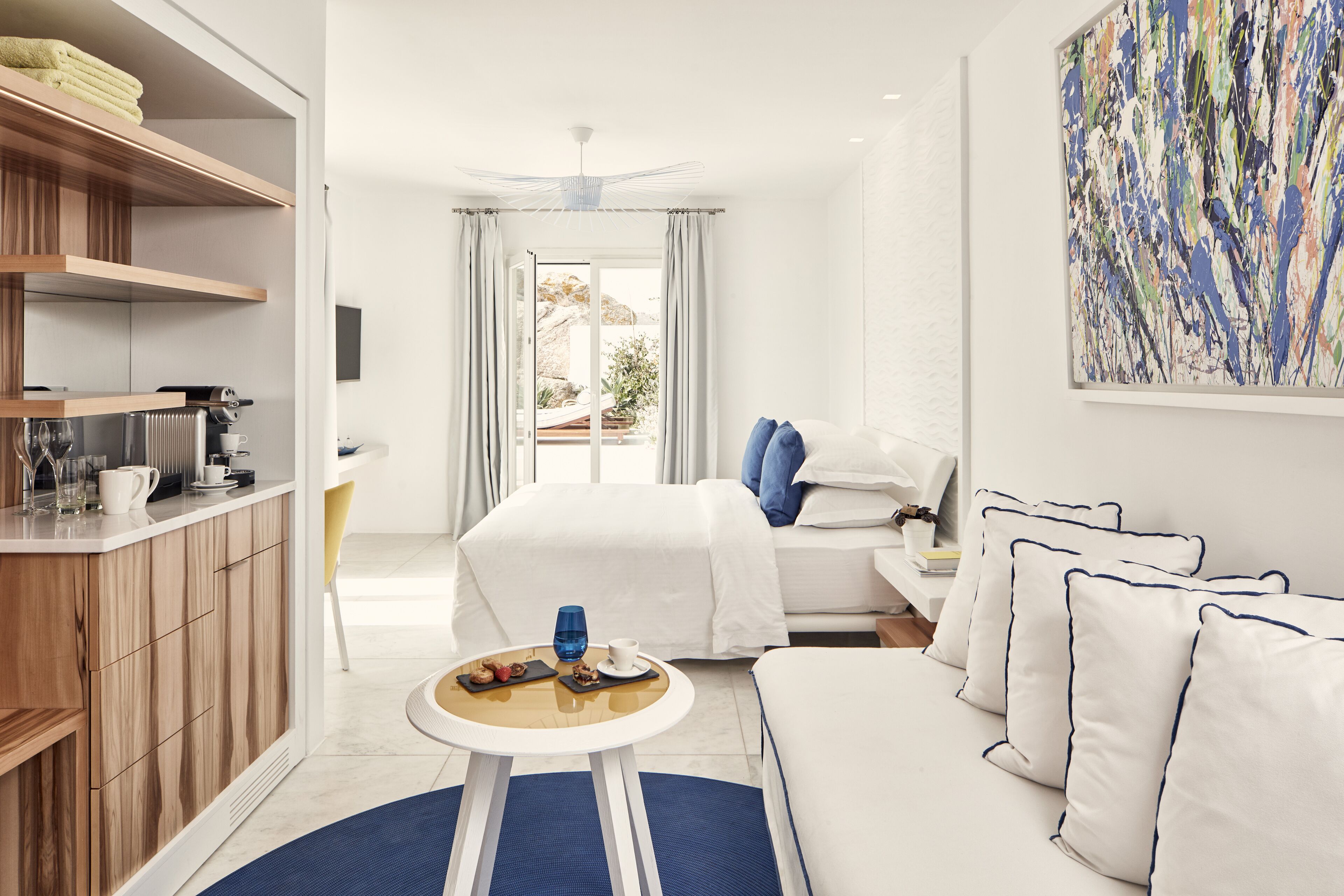 Een moderne hotelkamer met kitchenette, een bed met wit beddengoed, een blauw-wit kleurenschema en een groot abstract schilderij aan de muur.