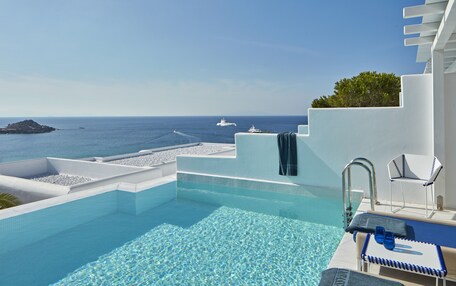 An infinity pool with clear blue water, a white infinity pool edge, and a view of the sea with boats.