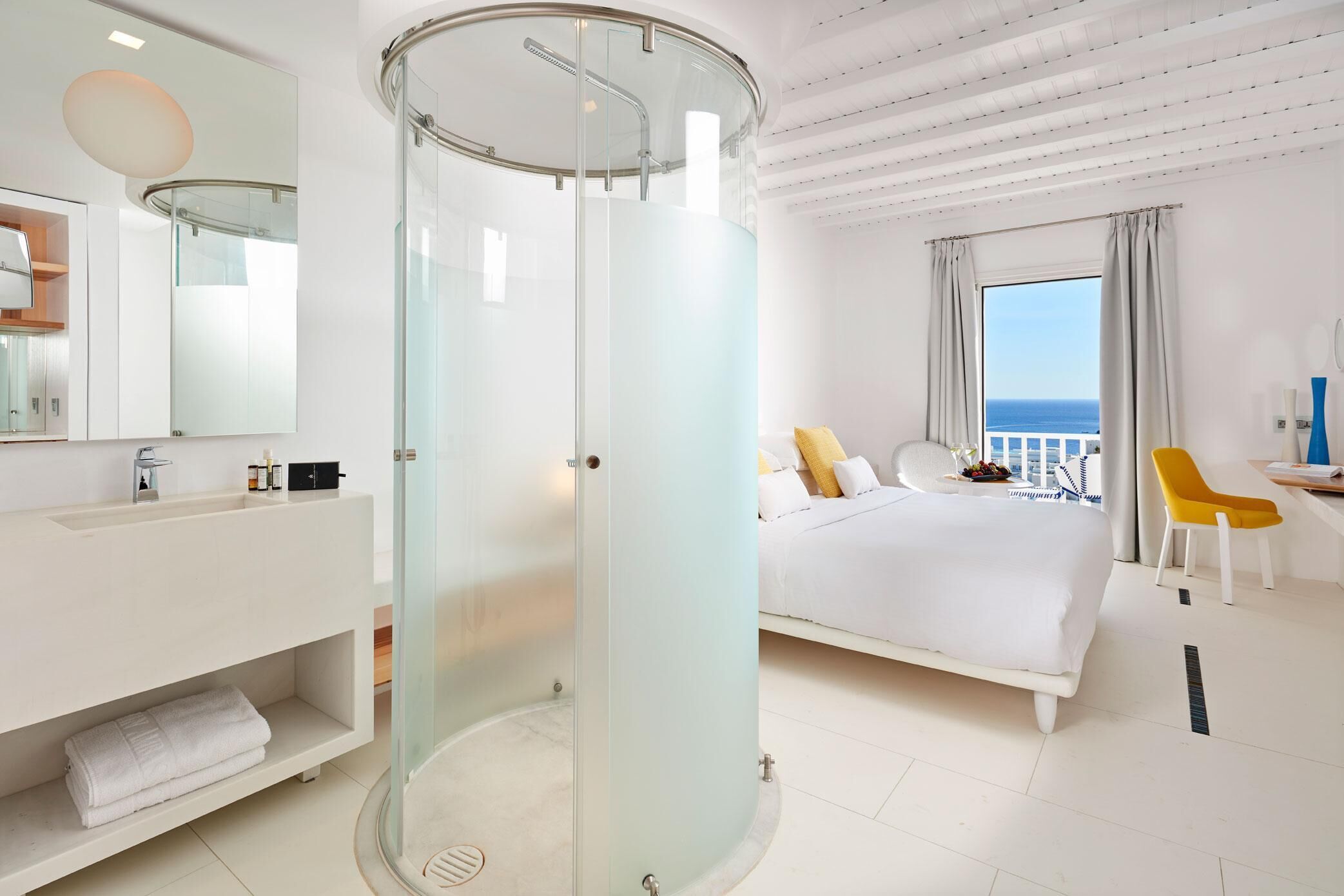 Une chambre d’hôtel moderne dotée d’une douche en verre, d’un lit blanc, d’une salle de bain avec un lavabo et un miroir, et offrant une vue sur la mer.