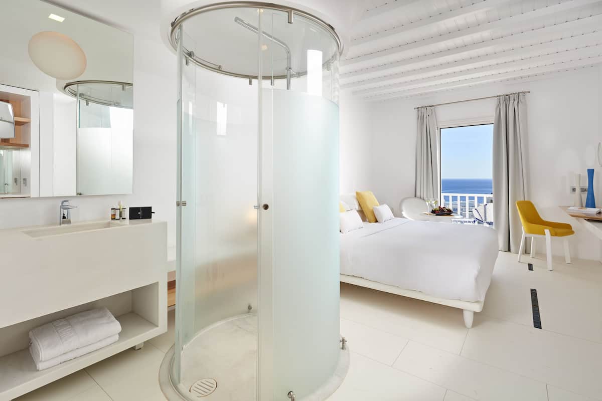 Een moderne hotelkamer met een douchecabine van glas, een wit bed, een gele stoel en uitzicht op zee.