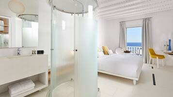 Una habitación de hotel moderna con ducha envuelta en vidrio, una cama blanca, una silla amarilla y vista al mar.