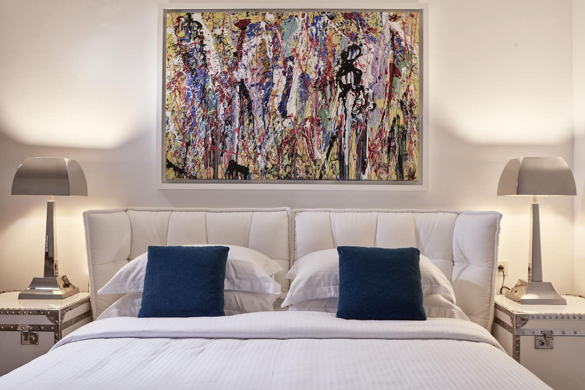 Een hotelkamer met een groot abstract schilderij aan de muur, een bed met witte lakens en twee blauwe kussens, en twee nachtlampen op bijpassende nachtkastjes.