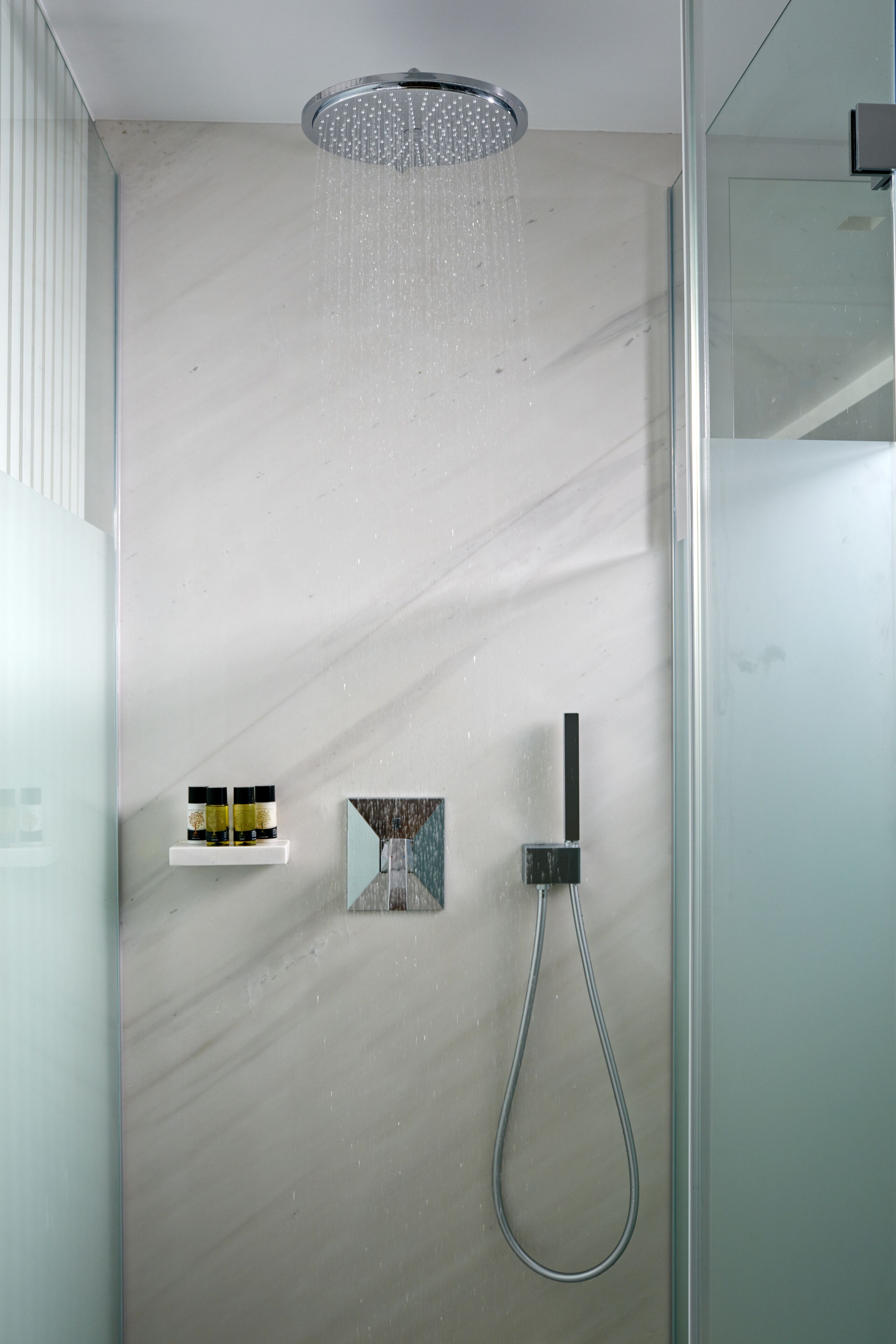 Une douche moderne dotée d’un pommeau de douche à pluie, d’un pommeau de douche à main et d’une petite étagère pour les articles de toilette.