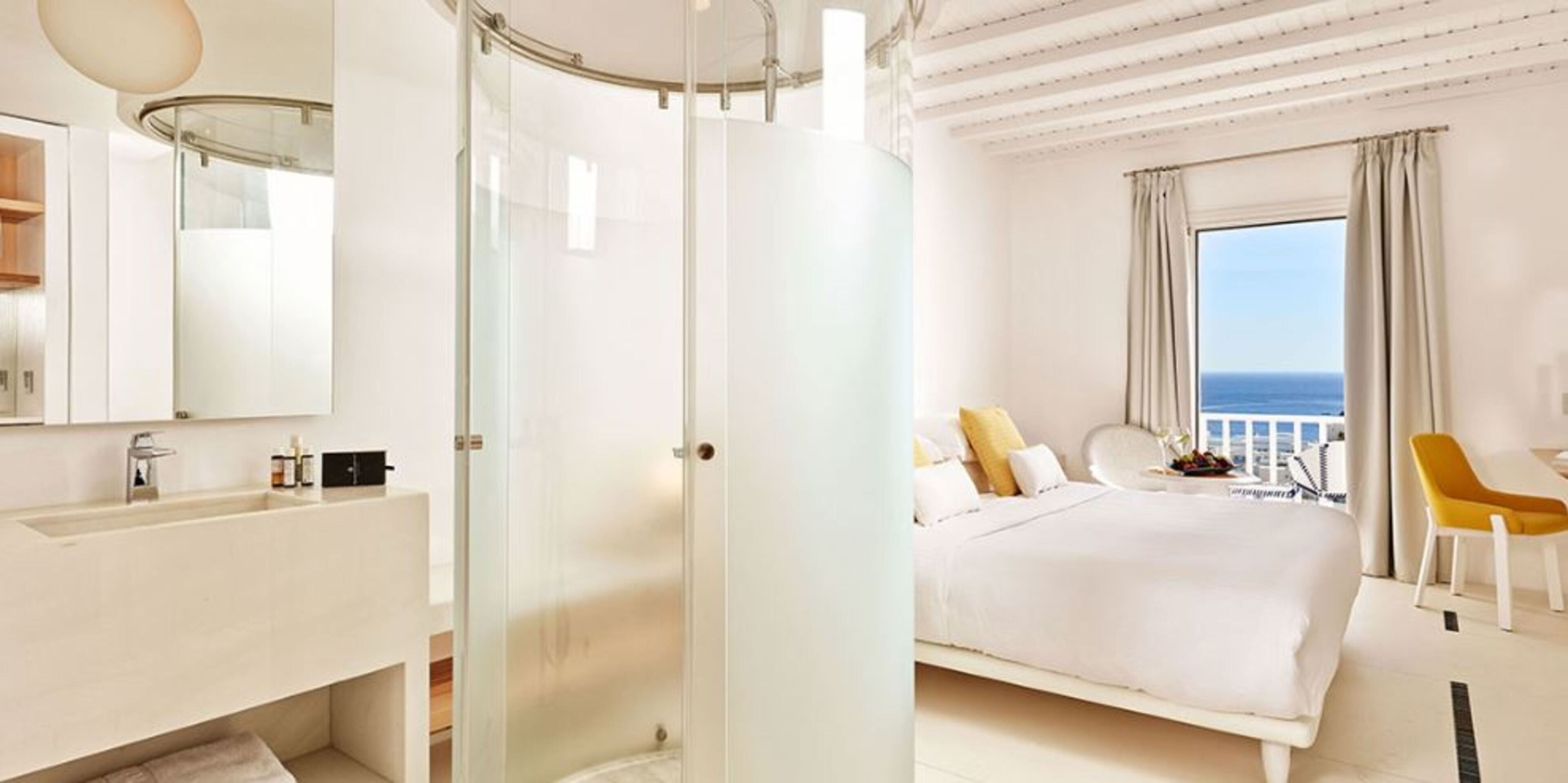 Une chambre d’hôtel moderne dotée d’une douche en verre, d’un grand lit, d’un balcon avec vue et d’une décoration aux tons blancs.