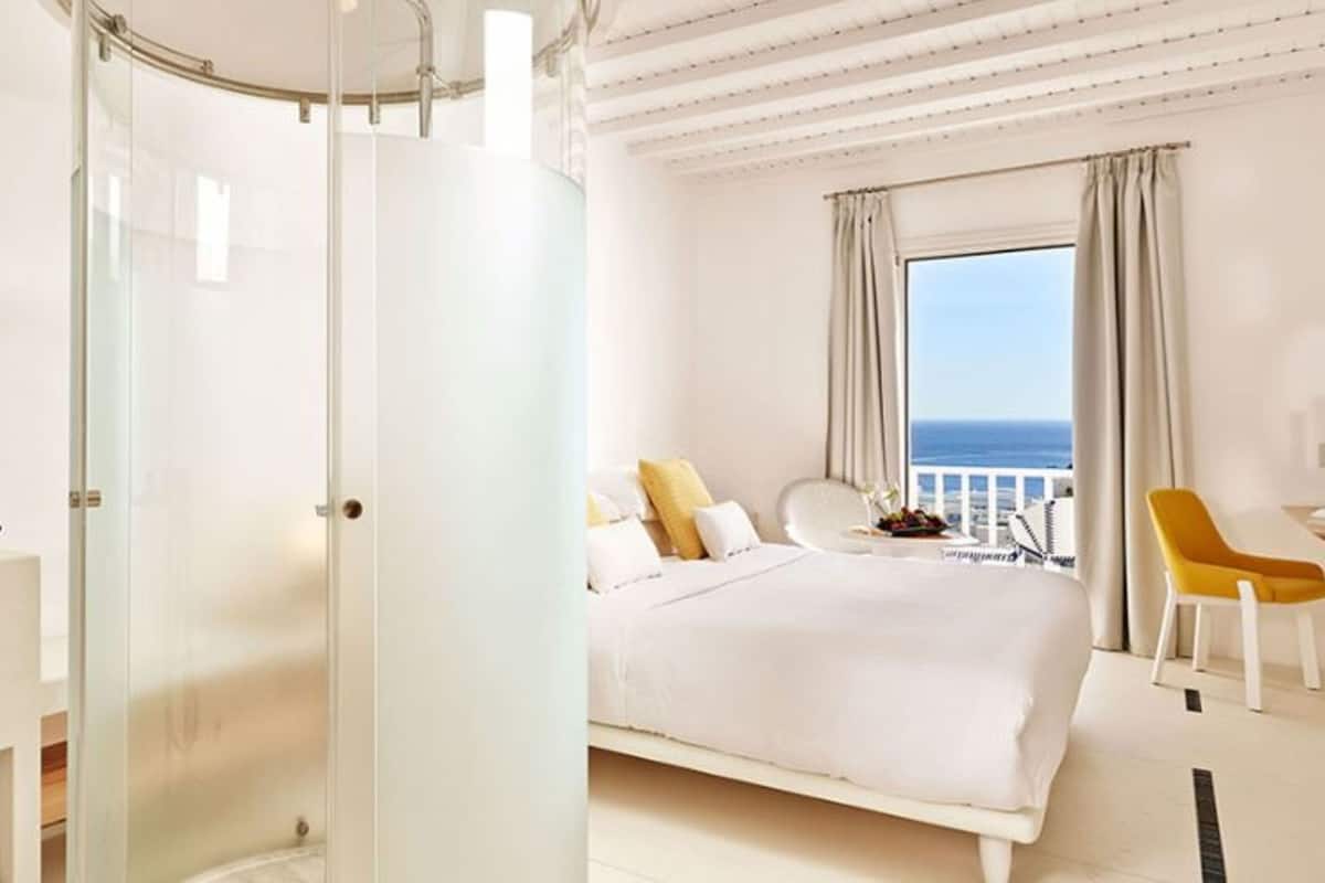 A modern hotel room with a glass-enclosed shower, a large bed, a balcony with a view, and a white color scheme.