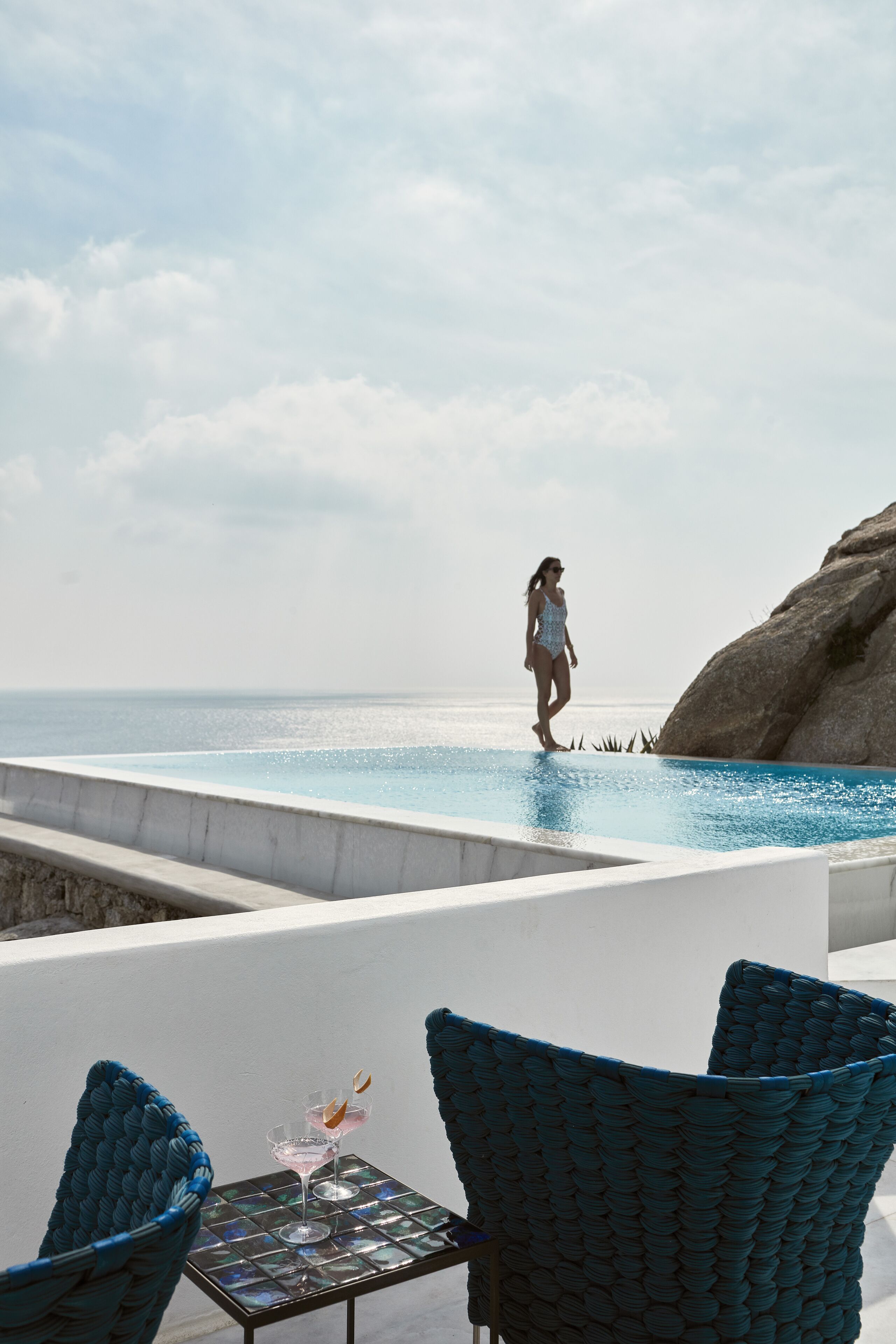 A poolside area with a person standing by the pool, two wicker chairs, and a table with a drink.