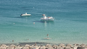 Ein Strand mit klarem türkisblauem Wasser, einem Boot und einer Person, die auf einem Paddleboard fährt.