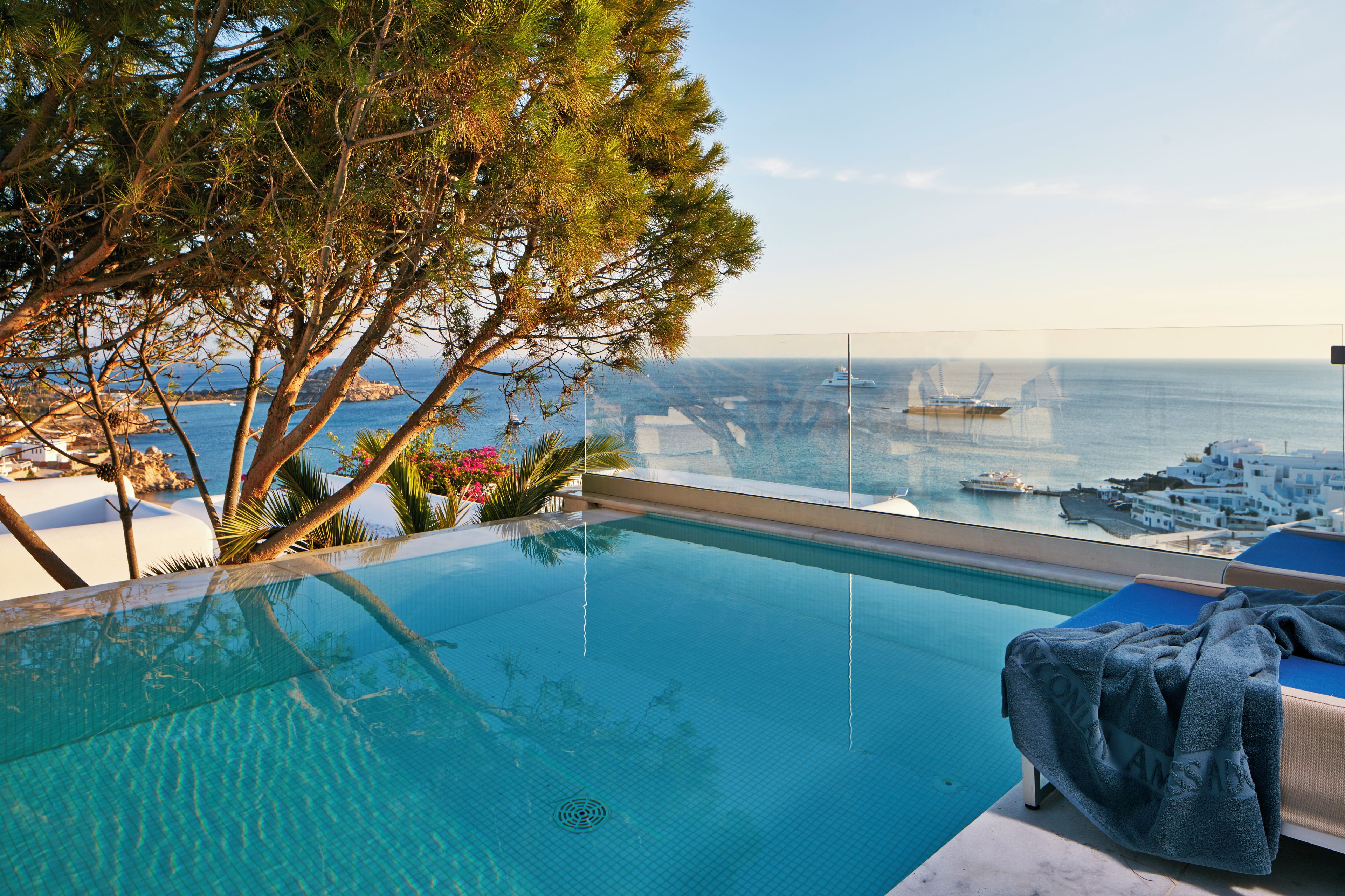 Una piscina con vista despejada al mar y a barcos, rodeada de árboles y un área de descanso con una toalla.