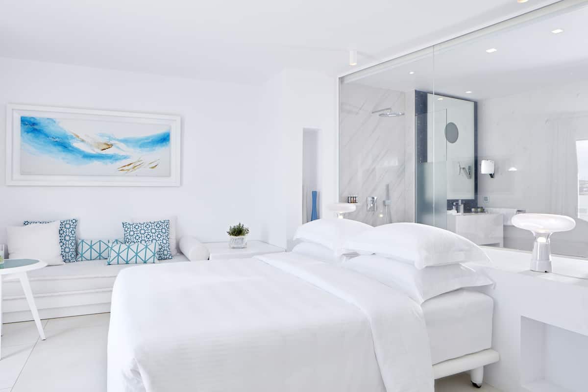 Een moderne hotelkamer met een groot bed, een nachtkastje, een bureau, een stoel, een bank met gestreepte kussens, een wanddecoratie, een grote spiegel en een badkamer met een wastafel en spiegel.