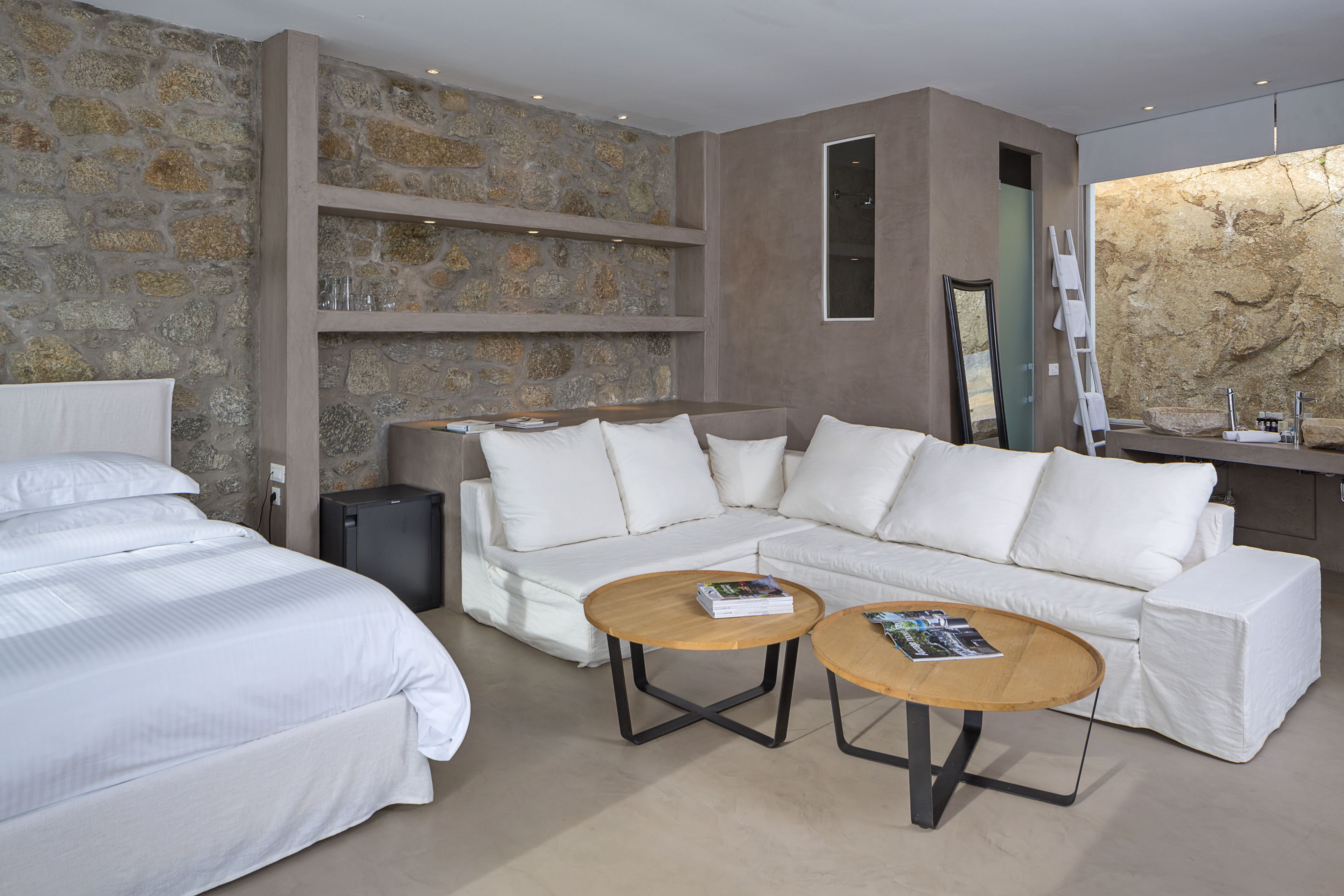 A modern hotel room with a stone accent wall, a white sofa, a bed, and two coffee tables.