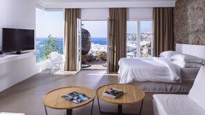 Ein modernes Hotelzimmer mit einem großen Bett, einer Sitzecke, zwei runden Holztischen, einem Fernseher und Blick auf das Meer und die Stadt.