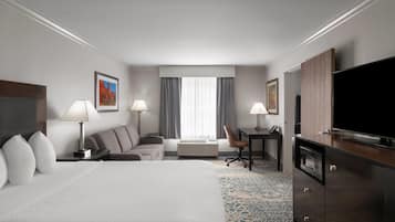 A hotel room with a large bed, two bedside tables with lamps, a desk with a chair, a flat-screen TV, and a large window with curtains.