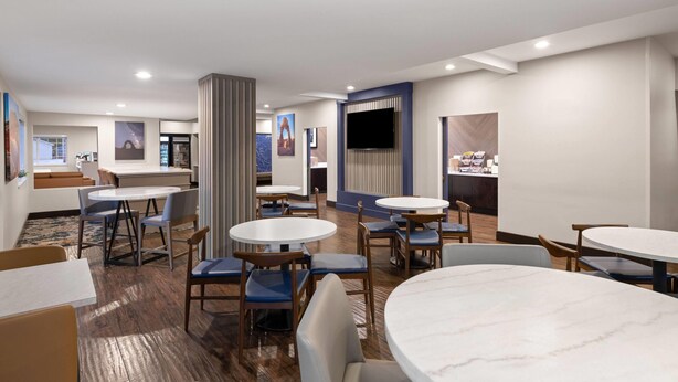 Un hall d’hôtel moderne avec des tables rondes, des fauteuils bleus rembourrés, un grand téléviseur à écran plat et un comptoir de réception.