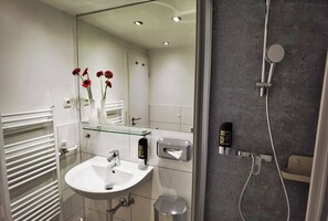 Un baño moderno con un lavamanos blanco, repisa de vidrio, secamanos automático, dispensador de toallas y ducha con cabezal de ducha manual.
