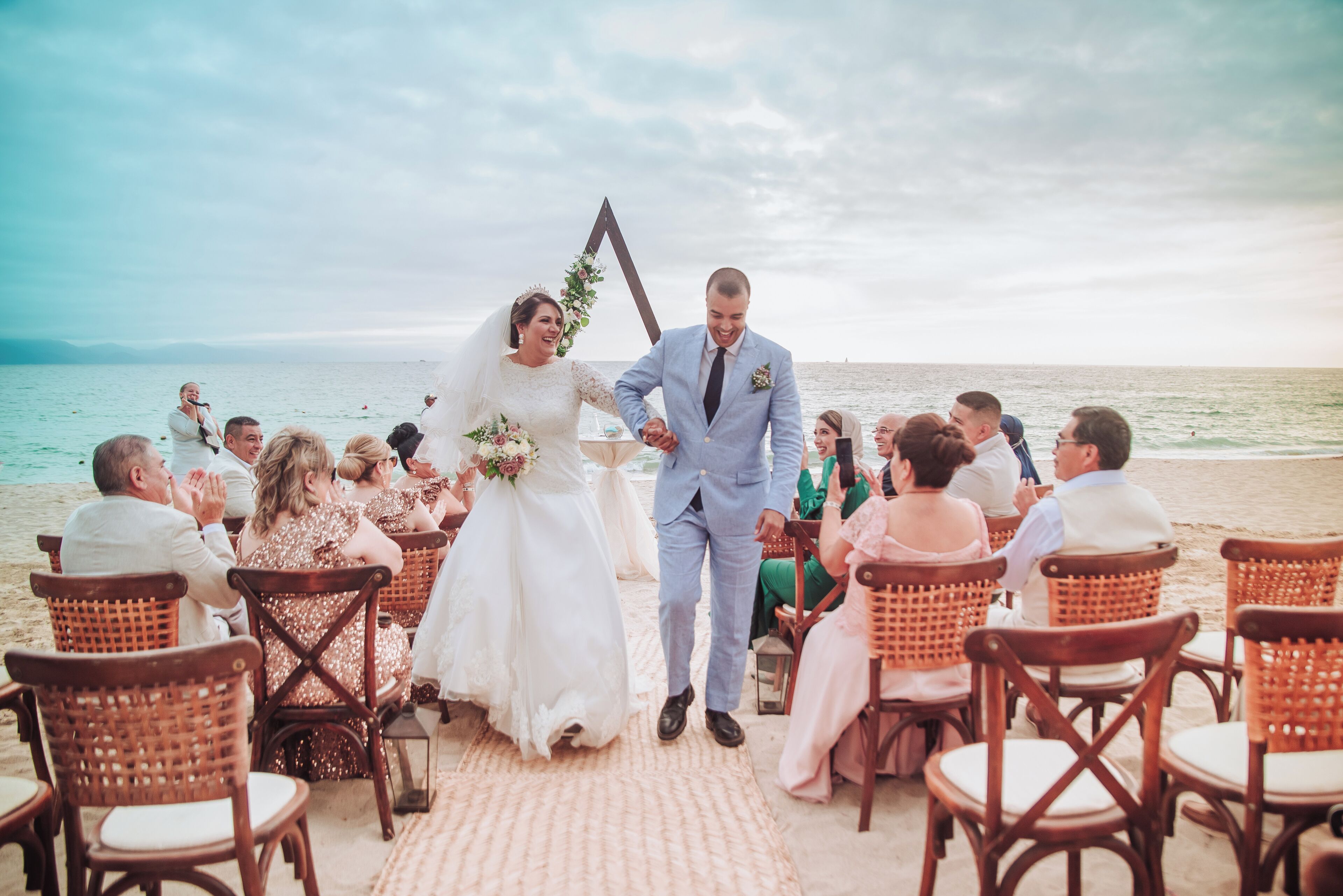 A wedding ceremony on a beach with guests seated on wicker chairs facing the aisle.