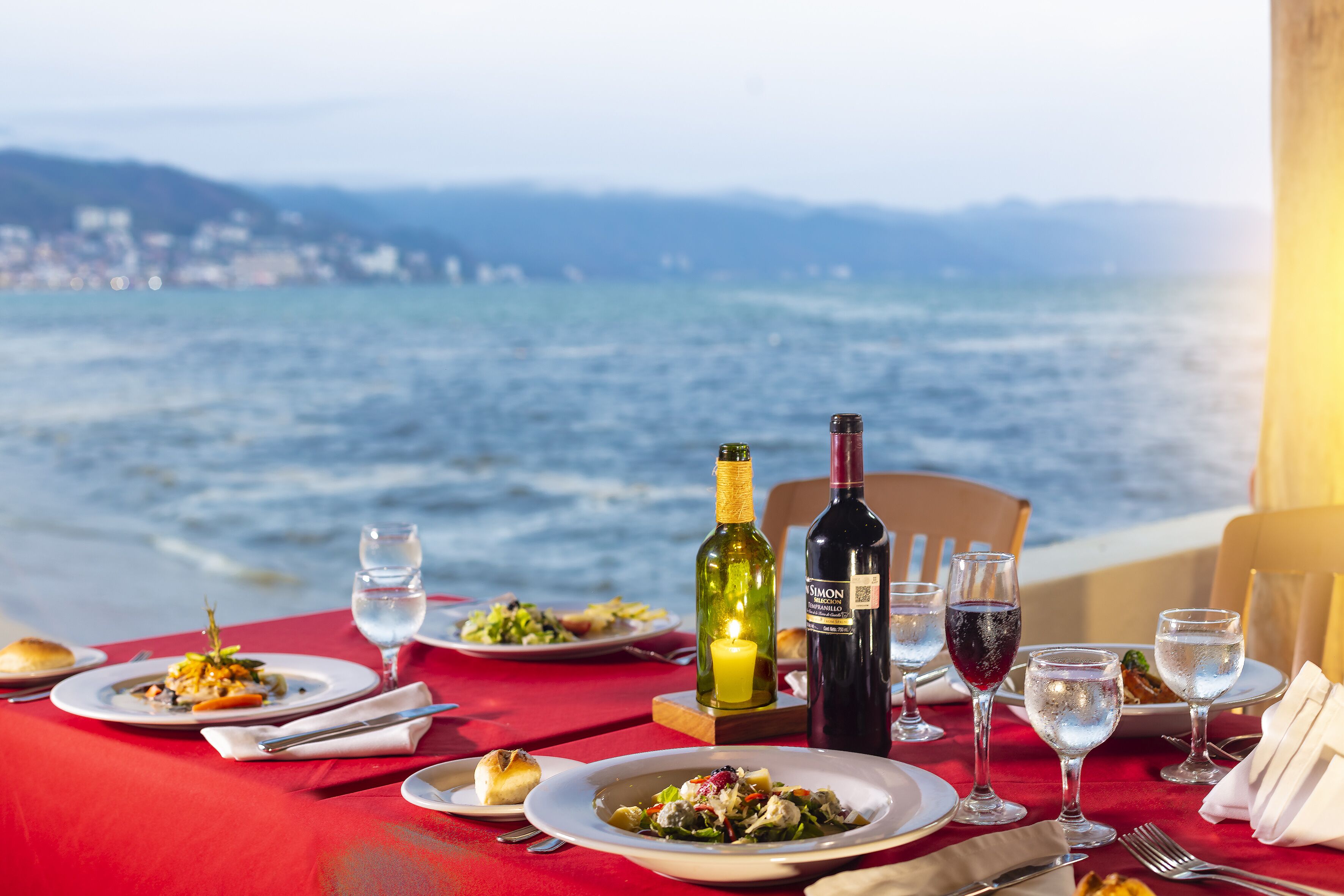 Mesa dispuesta con platos de comida, vasos y botellas de vino, con vista a una masa de agua y una costa lejana.