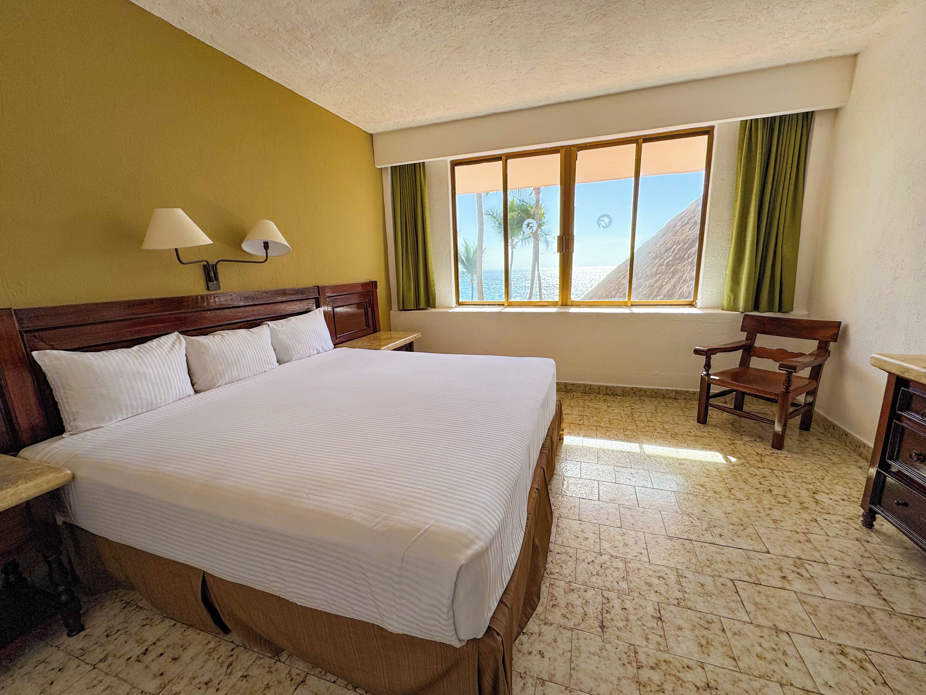 Habitación de hotel con una cama grande, dos lámparas de noche, una silla de madera, una mesita de noche y una ventana con vista a la playa.
