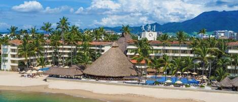 Un complejo turístico en la playa con estructuras de techo de paja, palmeras y una zona de piscina.