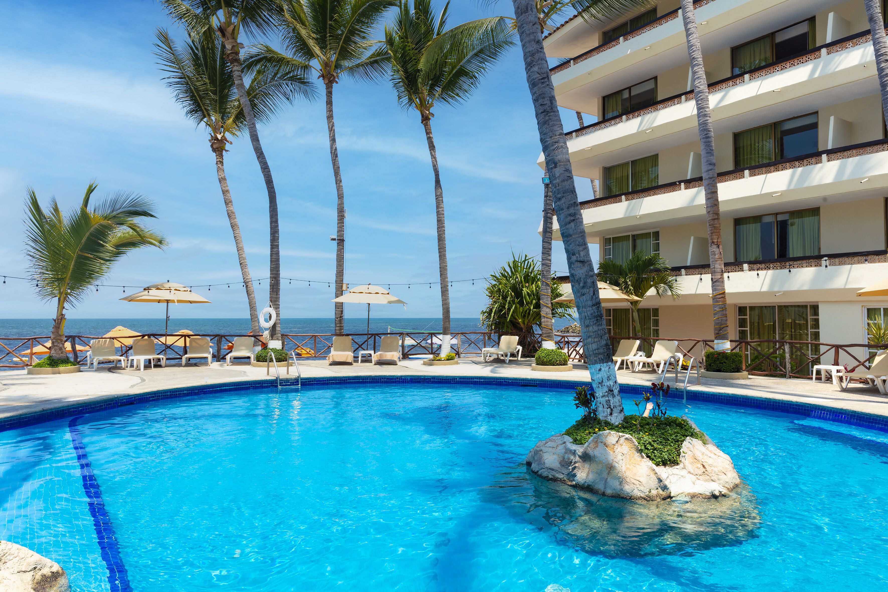 Una piscina con agua azul cristalina, rodeada de palmeras y sillones de descanso, con vista al océano y un edificio de varios pisos al fondo.